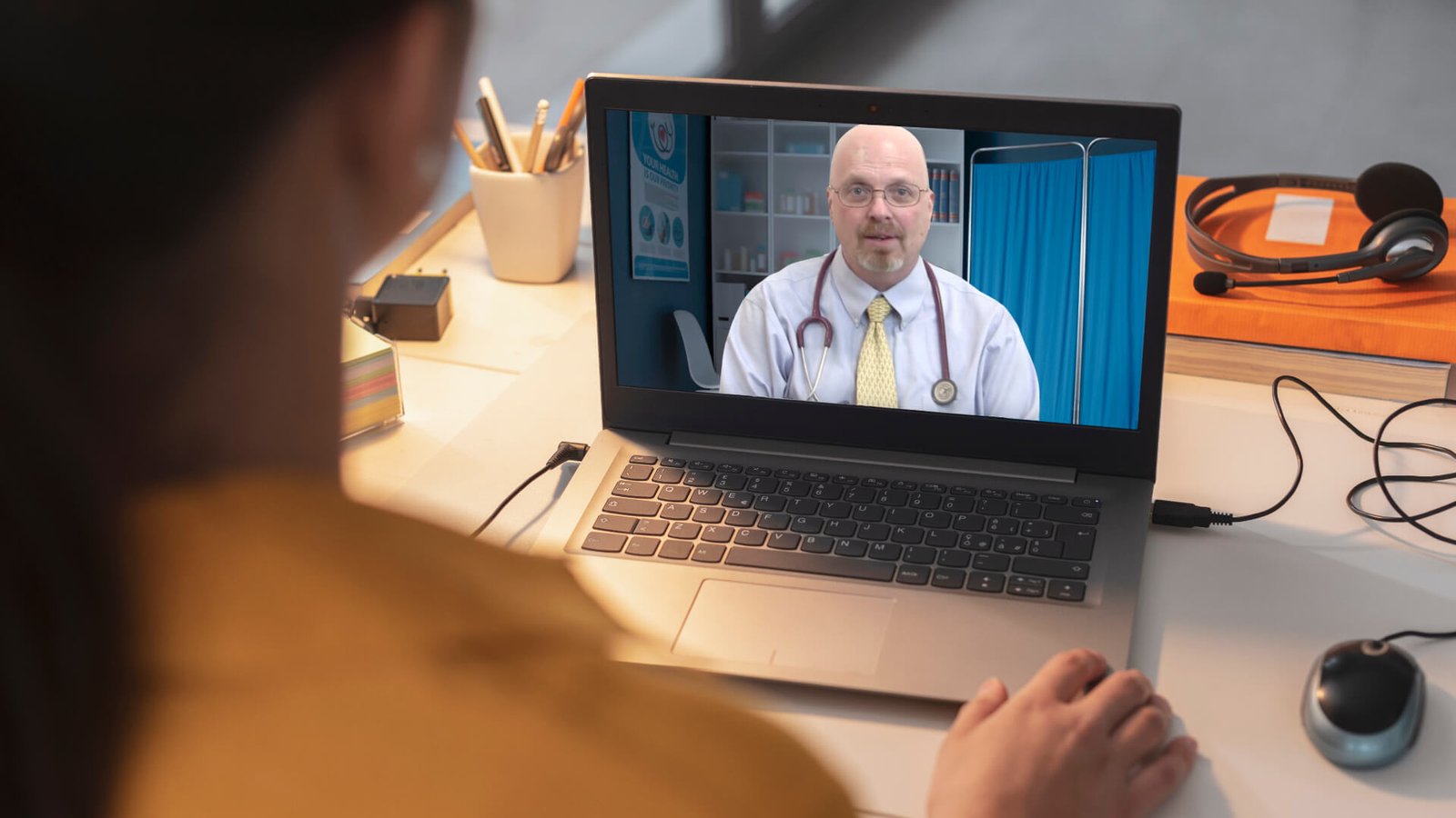 the outline of a patients shoulder sitting on the laptop speaking with a medical professional wearing a shirt and stethoscope around his neck